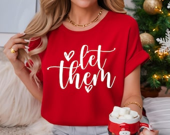 May include: A red t-shirt with the white text "let them" in a cursive font, with heart accents. The shirt is a crew neck style. A mug with marshmallows is visible in the image.