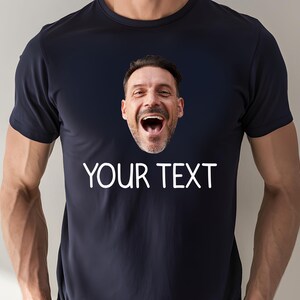 May include: Navy blue t-shirt with a printed image of a laughing man's face. The text "YOUR TEXT" is printed in white below the image. The shirt has short sleeves and a crew neck.