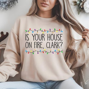 May include: A beige sweatshirt with a Christmas lights design and the text "Is your house on fire, Clark?"