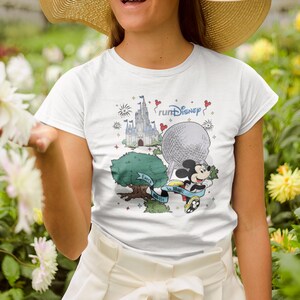 May include: White t-shirt with a graphic print of Mickey Mouse running through a cartoon version of Walt Disney World. The graphic includes the text "runDisney" and "Every Mile a Magic Moment".