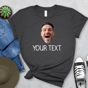 May include: Dark gray t-shirt with a printed image of a man's face and the text "YOUR TEXT" in white. The shirt is styled with blue jeans, a hat, a cactus, and black and white sneakers.