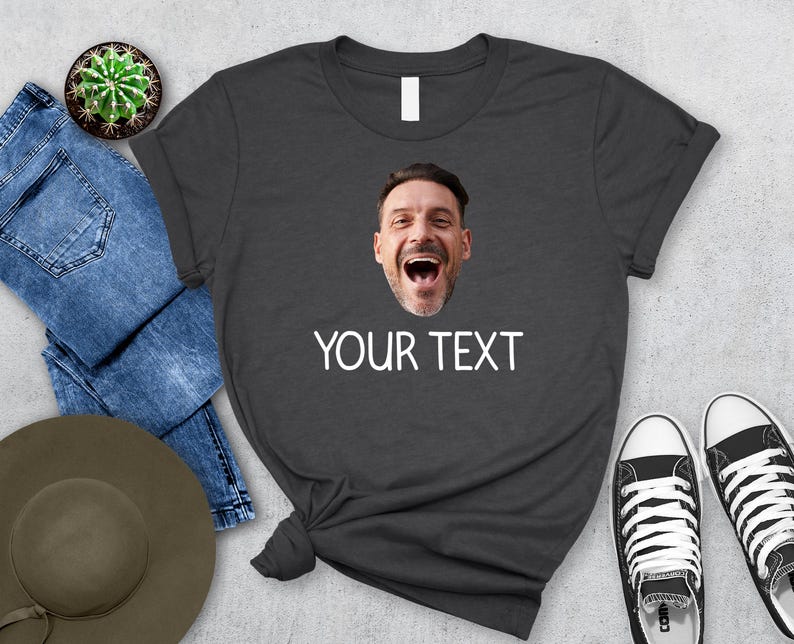 May include: Dark gray t-shirt with a printed image of a man's face and the text "YOUR TEXT" in white. The shirt is styled with blue jeans, a hat, a cactus, and black and white sneakers.