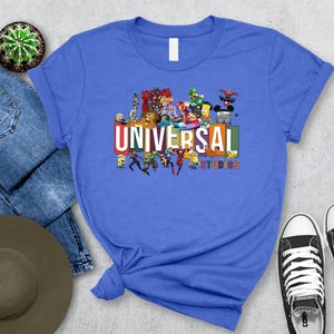 May include: A blue t-shirt featuring the text "UNIVERSAL STUDIOS" in a colorful block design. The shirt is adorned with cartoon characters from various Universal Studios franchises. A pair of black and white sneakers are visible.
