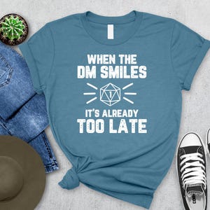 May include: Teal t-shirt with the text "WHEN THE DM SMILES IT'S ALREADY TOO LATE" in white. The shirt features a graphic of a D20 die. The shirt is paired with blue jeans, a hat, a cactus, and black and white sneakers.