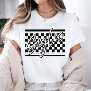 May include: White t-shirt featuring the word "Eagles" in a stylized script font. The word is set against a black and white checkered background with horizontal black lines above and below. The shirt has short sleeves.