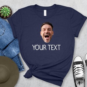 May include: Navy blue t-shirt with a printed image of a man's face and the text "YOUR TEXT" in white. The shirt is paired with blue jeans, a hat, a cactus, and black and white sneakers.