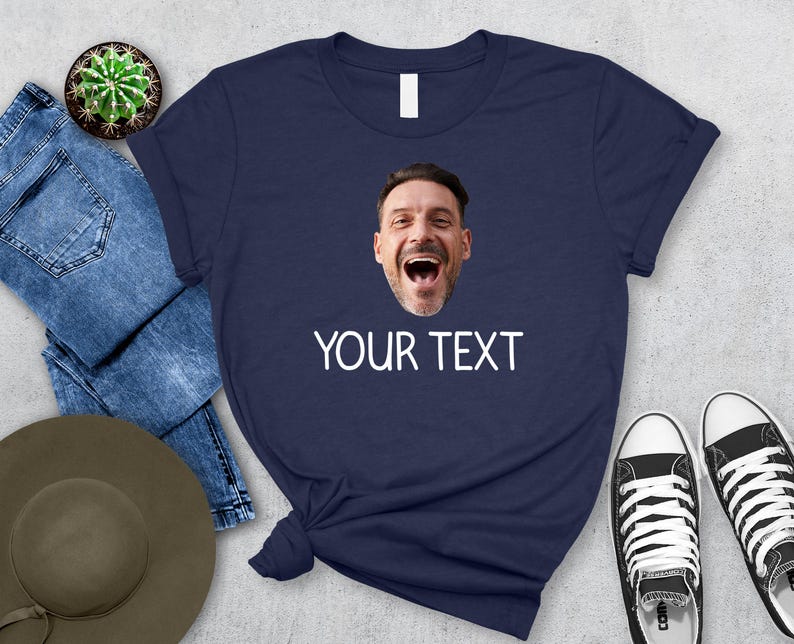 May include: Navy blue t-shirt with a printed image of a man's face and the text "YOUR TEXT" in white. The shirt is paired with blue jeans, a hat, a cactus, and black and white sneakers.