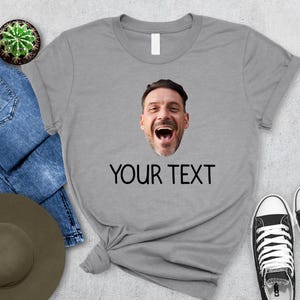 May include: A gray t-shirt featuring a smiling face and the text "YOUR TEXT" in black. The shirt is paired with blue jeans, a hat, and black and white sneakers. A small cactus in a pot is also visible.