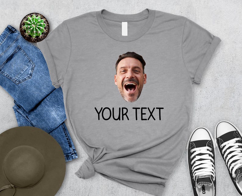May include: A gray t-shirt featuring a smiling face and the text "YOUR TEXT" in black. The shirt is paired with blue jeans, a hat, and black and white sneakers. A small cactus in a pot is also visible.