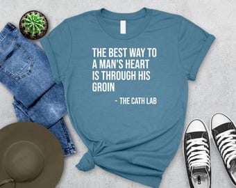 May include: Teal t-shirt with the text "THE BEST WAY TO A MAN'S HEART IS THROUGH HIS GROIN - THE CATH LAB" in white. The shirt is styled with blue jeans, a hat, a cactus, and black and white sneakers.