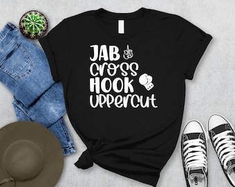 May include: Black t-shirt featuring the text "JAB CROSS HOOK UPPERCUT" in white, accompanied by boxing-themed illustrations. The shirt is shown with blue denim jeans, a hat, a cactus, and black and white trainers.
