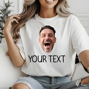 May include: White t-shirt featuring a smiling man's face and the text "YOUR TEXT" in black. The shirt is a casual style, suitable for everyday wear. The design is simple and bold.