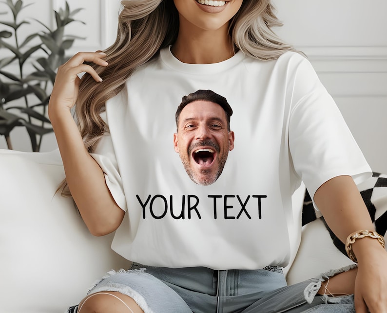 May include: White t-shirt featuring a smiling man's face and the text "YOUR TEXT" in black. The shirt is a casual style, suitable for everyday wear. The design is simple and bold.
