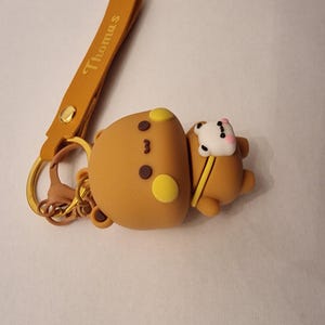 May include: A brown bear keychain with a yellow strap that reads "Thomas". The bear has yellow accents and a small white bear companion. The keychain is attached to a gold-colored ring.