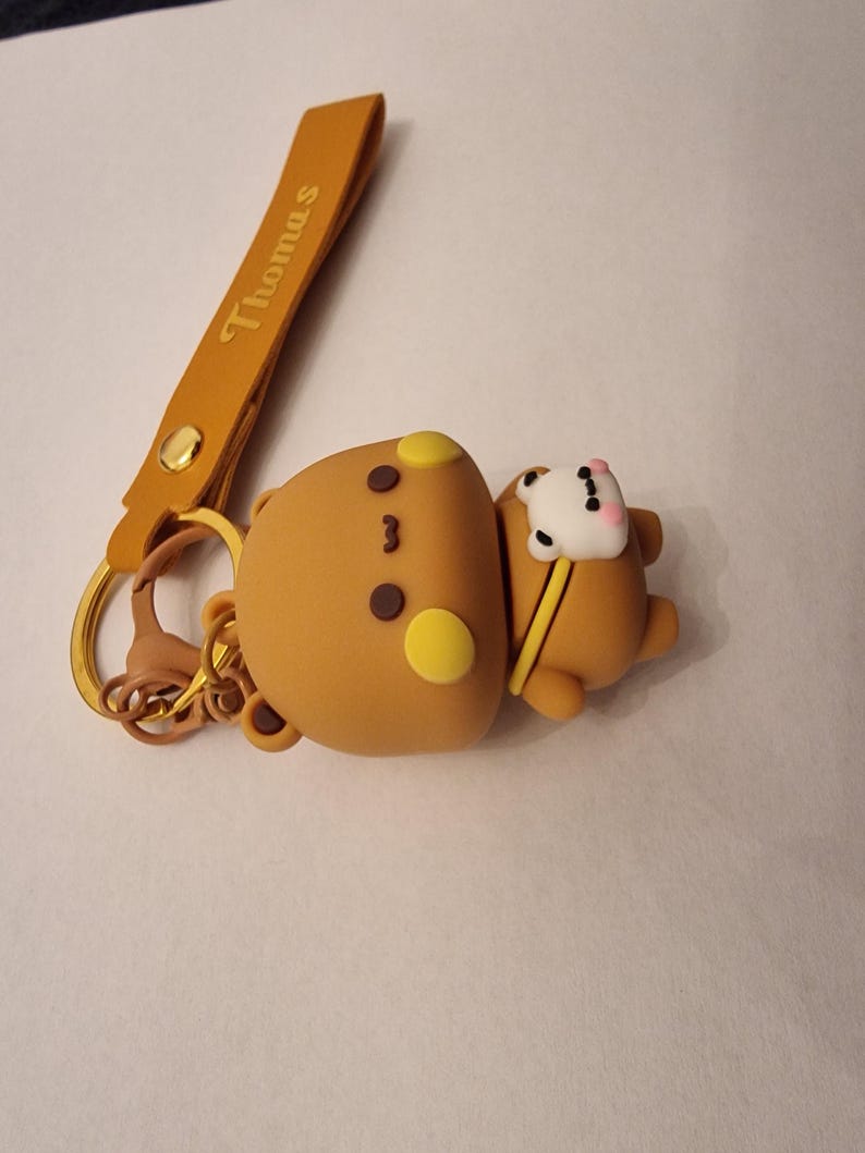 May include: A brown bear keychain with a yellow strap that reads "Thomas". The bear has yellow accents and a small white bear companion. The keychain is attached to a gold-colored ring.