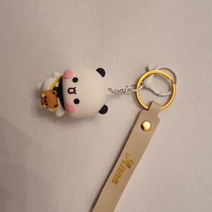 May include: A white and black panda keychain with a small brown bear accessory. The keychain has a gold-colored ring and a beige leather strap with the name "Anna" in gold lettering. The panda has pink cheeks.