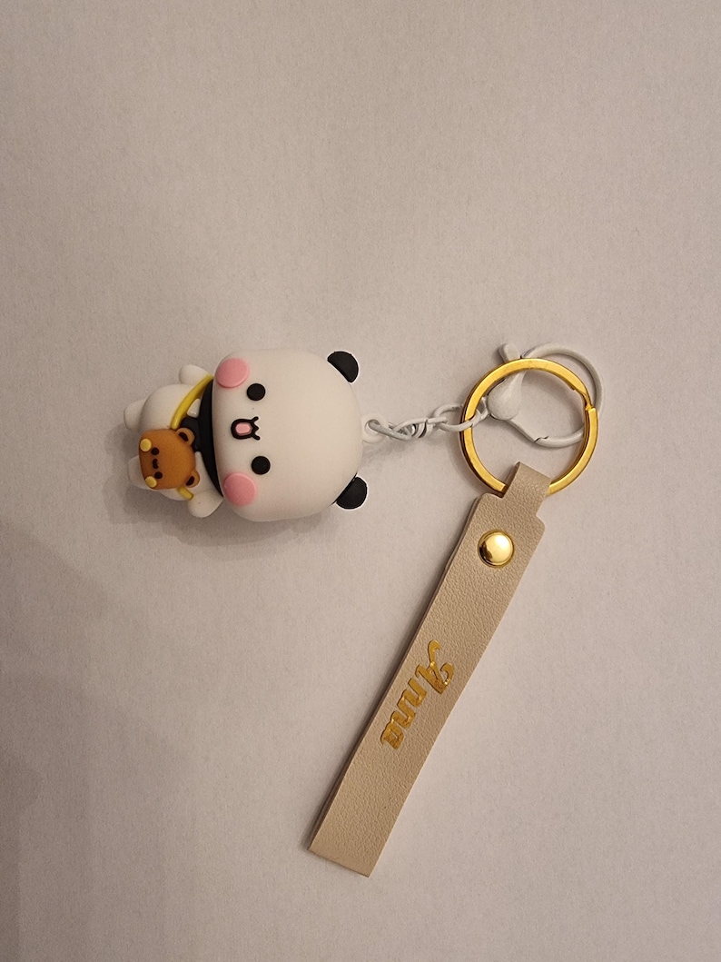 May include: A white and black panda keychain with a small brown bear accessory. The keychain has a gold-colored ring and a beige leather strap with the name "Anna" in gold lettering. The panda has pink cheeks.