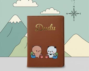 May include: Brown passport holder with the gold script text "Dudu". The cover features two cartoon bears with luggage, set against a travel-themed background with mountains, clouds, a compass, and an airplane.