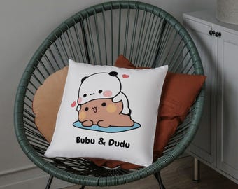 May include: White square pillow with a cartoon illustration of two pandas, one on top of the other, with the text "Bubu & Dudu" in black. The pillow is on a green chair with orange and tan pillows.