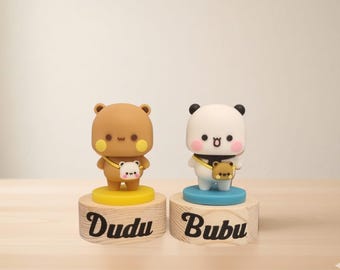 May include: Two small figurines on wooden bases. The left figurine is a brown bear with yellow cheeks and a small white bag, labeled Dudu. The right figurine is a white panda with pink cheeks and a small brown bag, labeled Bubu. Both have round heads.