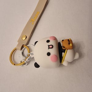May include: A white panda keychain with black accents, pink cheeks, and a small brown bear attached. The keychain has a gold ring and a beige strap with the name "Anna" in gold lettering.