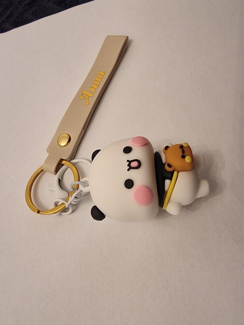 May include: A white panda keychain with black accents, pink cheeks, and a small brown bear attached. The keychain has a gold ring and a beige strap with the name "Anna" in gold lettering.