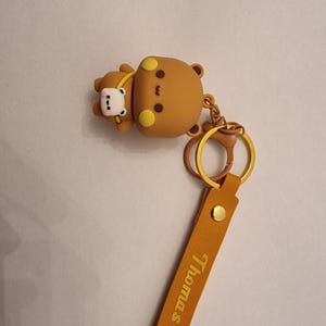 May include: A brown bear keychain with a smaller bear attached. The keychain has a gold-colored ring and a tan leather strap with the name "Thomas" printed on it. The bears have yellow accents.