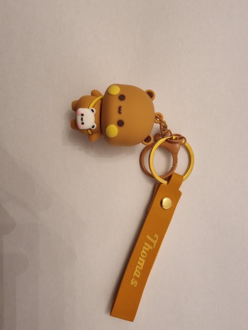 May include: A brown bear keychain with a smaller bear attached. The keychain has a gold-colored ring and a tan leather strap with the name "Thomas" printed on it. The bears have yellow accents.