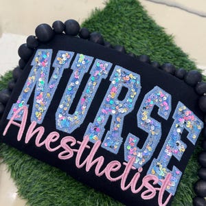 Könnte beinhalten: Schwarzer Pullover mit den Worten "NURSE Anesthetist" in blauen und rosa Pailletten.