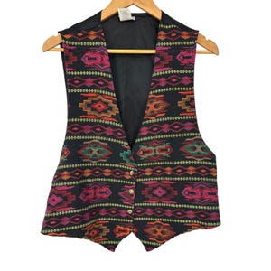 May include: A black vest featuring a vibrant, multi-colored geometric pattern in shades of pink, orange, green, and brown. The garment has a V-neckline, a solid black interior lining, and three small, circular metallic buttons along the front closure.