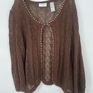 Può includere: Un cardigan marrone con apertura frontale e un delicato design a maglia traforata. Il maglione ha un orlo smerlato ed è impreziosito da piccole perline colorate lungo lo scollo e i bordi anteriori. L'etichetta del marchio è visibile.