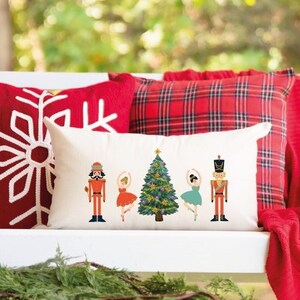 Może przedstawiać: Biały huśtawka werandowa z czerwonymi i białymi poduszkami w kratę i białą poduszką z motywem choinki i orzechów. Huśtawka jest ozdobiona czerwonym kocem i zielonym wieńcem.