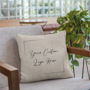 May include: A beige throw pillow with a black border and the text "Your Custom Logo Here" in a black script font.