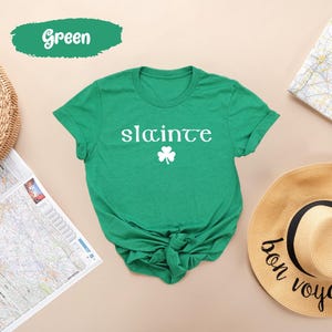 May include: A green t-shirt with the word "sláinte" in white, and a white shamrock. The shirt is knotted at the waist. A green banner above the shirt says "Green". A straw hat and map are in the background.