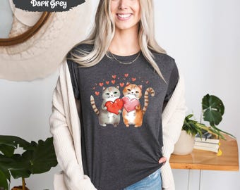 May include: A heather dark grey t-shirt featuring a graphic of two cats holding red hearts. The cats are a grey tabby and an orange tabby. The graphic is surrounded by small red hearts.