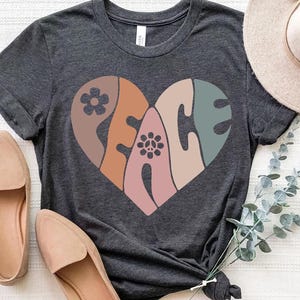 Könnte beinhalten: Ein dunkelgraues T-Shirt mit einem Peace-Zeichen in Herzform. Das Peace-Zeichen besteht aus verschiedenen farbigen Abschnitten, darunter Braun, Orange, Rosa und Grün. Das Peace-Zeichen hat ein Blumenmuster in der Mitte.
