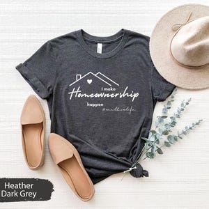 Może przedstawiać: Szary melanżowy t-shirt z nadrukiem "I make Homeownership happen #realestatelife" w kolorze białym. T-shirt przedstawia grafikę domu z sercem.