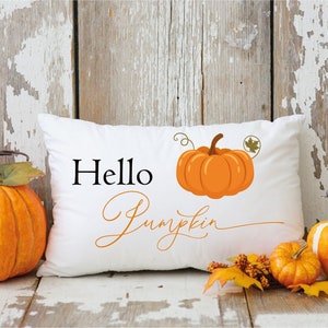 May include: White cushion with the text "Hello Pumpkin" in black and orange lettering. A small orange pumpkin with a green stem and a brown leaf is on the cushion. The cushion is sitting on a wooden surface with two small pumpkins and autumn foliage.