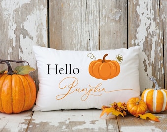 May include: White throw pillow with the text "Hello Pumpkin" in black and orange lettering. A small orange pumpkin with a green stem and a brown leaf is on the pillow. The pillow is sitting on a wooden surface with two small pumpkins and fall foliage.
