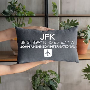 Könnte beinhalten: Ein graues Kissen mit weißem Text, der "JFK 38 51' 8.99° N 40 63' 6.71° W JOHN F. KENNEDY INTERNATIONAL" lautet, und einem weißen Flugzeugsymbol.