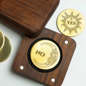Puede incluir: Una caja de adivinación de madera con monedas de latón. Una moneda dice "NO" con una luna creciente, la otra "SÍ" con un sol. La caja es de madera oscura. Las monedas miden unos 5 cm de diámetro.