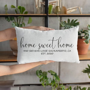 Puede incluir: Un cojín rectangular blanco con las palabras "home sweet home" en escritura negra. Debajo, la dirección "430 SIEVENS LANE SACRAMEBTO, CA EST. 2020". El cojín se sostiene frente a una estantería con plantas.