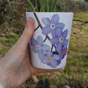May include: A white ceramic flower pot with a design of purple forget-me-not flowers.