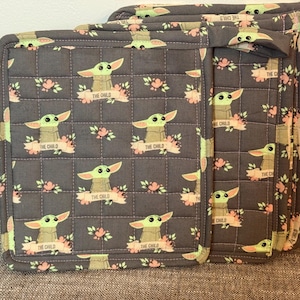 May include: Grey quilted pot holders with a repeating pattern of a green alien character with large ears and the text "THE CHILD" on a banner, along with pink floral accents. The pot holders are square and have a quilted design.