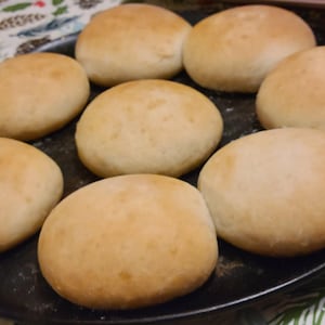 May include: A close-up of eight round, white bread rolls on a black baking sheet. The rolls are slightly browned and have a soft, fluffy texture.