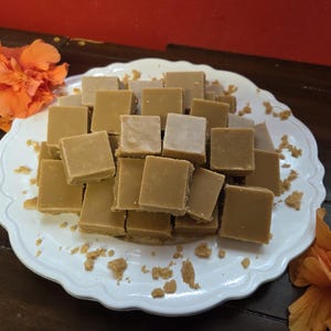 May include: A white, scalloped-edged plate piled high with square pieces of light brown fudge. The fudge is arranged in a slightly haphazard stack. Orange flowers are placed on either side of the plate, adding a pop of color.