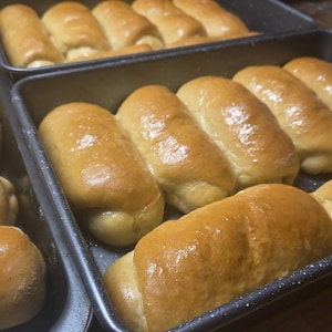 May include: Several baking pans filled with golden-brown, freshly baked rolls. The rolls are arranged in rows, with a glossy, glazed appearance. The image showcases a close-up view of the baked goods, highlighting their texture and color. The pans are dark-colored.