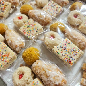 May include: An assortment of individually wrapped baked goods, including square frosted cookies with rainbow sprinkles, round cookies with red jelly centers, and golden-yellow, spherical treats. Some are glazed and others are dusted with powdered sugar.