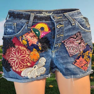 May include: Blue denim shorts featuring a button-fly closure and the Universal Thread brand label. The shorts are decorated with various colorful fabric patches, including floral and paisley patterns, black lace accents, and a silver butterfly.