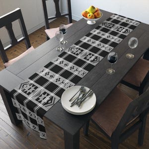 May include: A black and white patterned table runner with a geometric design is displayed on a dark wooden table. The table is set with wine glasses, a plate, and silverware. A bowl of fruit sits at the top.